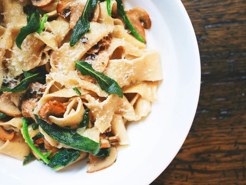 A white bowl of pappardelle with mushrooms, sage and grated parmesan on a dark timber table.